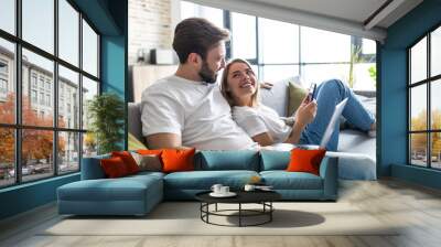 Beautiful young couple relaxing on a couch at home using laptop computer and mobile phone Wall mural